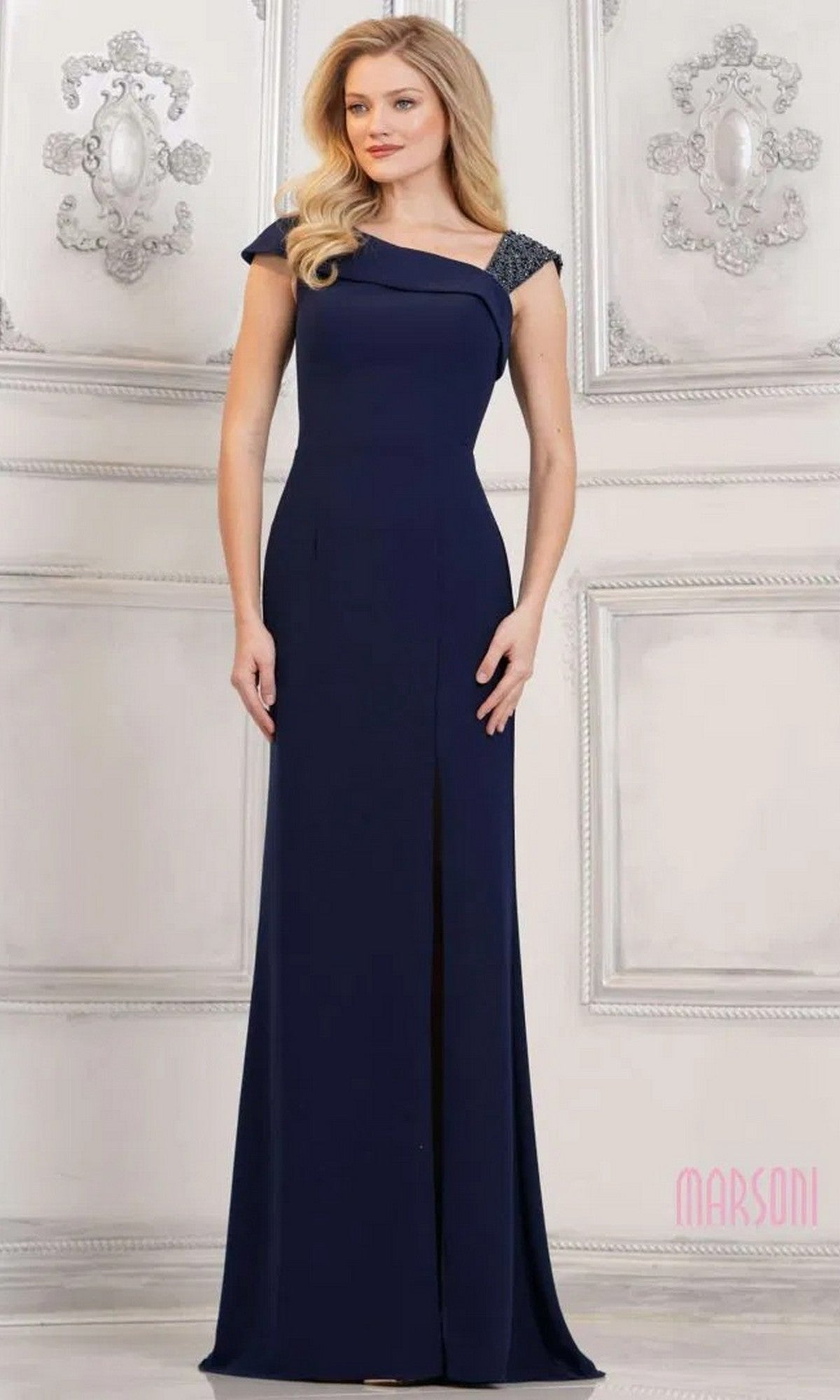 A woman stands in an elegant room, wearing the navy Long Formal Dress: Marsoni MV1296—a floor-length crepe gown with beaded detailing on one shoulder.